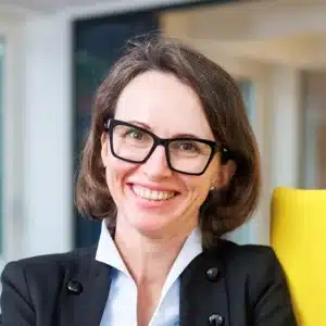 Porträt einer lächelnden Frau mit Brille und Blazer.